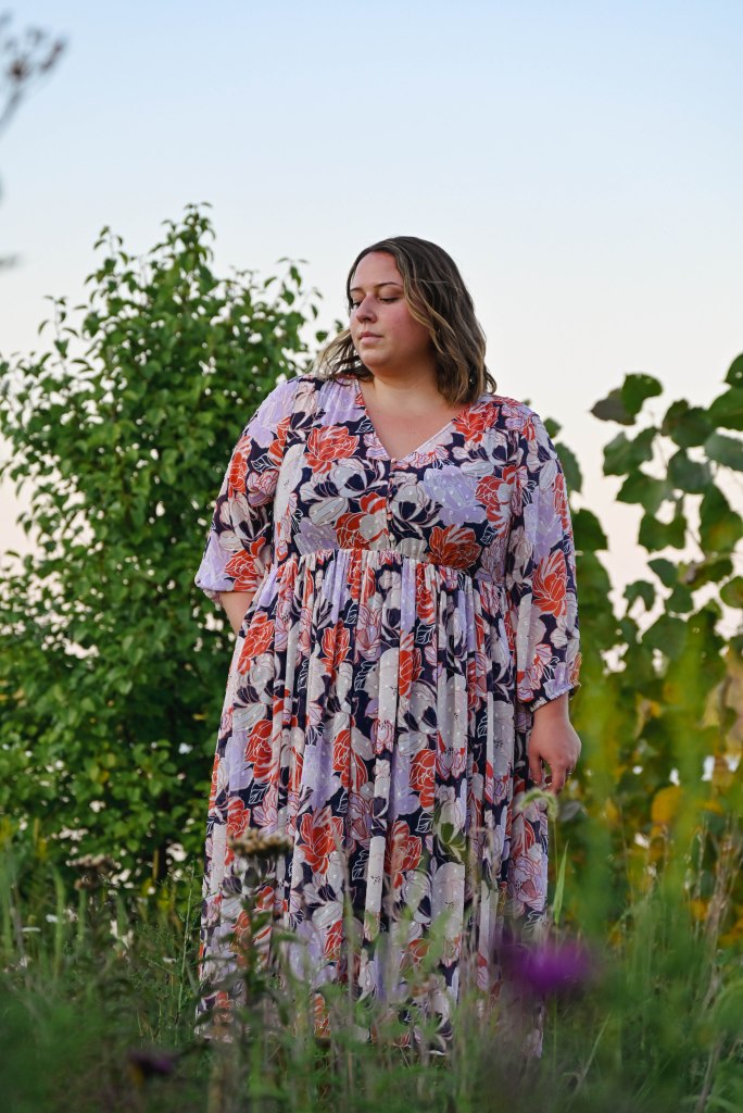 A woman stands in a purple and pink floral dress amongst green bushes and purple flowers. She has a soft expression, looking as though she's taking in the moment.