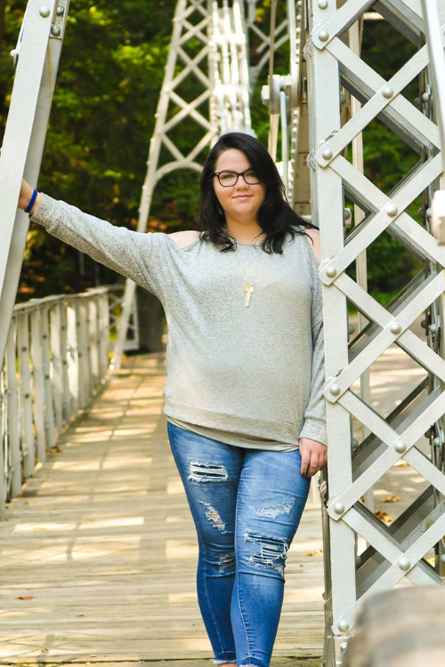 A soon-to-be graduate smiles softly at the camera while leaning against a silver framed bridge.